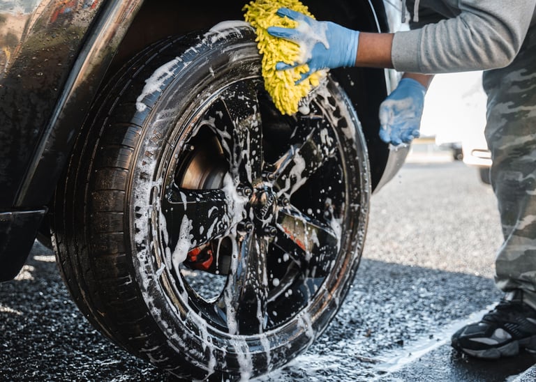 Person washing a car wheel with a yellow sponge and soapy water, wearing blue gloves